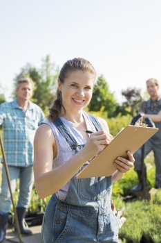 Portrait of beautiful supervisor holding clipboard with gardeners standing in background at plant nursery