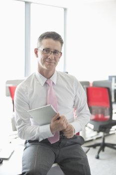 Portrait of confident businessman with tablet PC sitting on desk in office