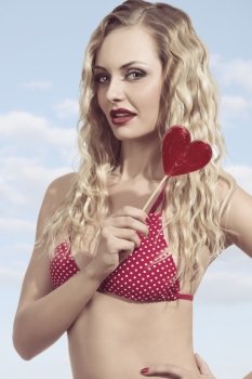 summer woman with long curly blonde hair wearing sexy bikini and eating red heart shaped lollipop. Funny shoot
