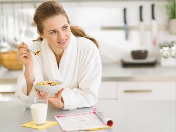 Thoughtful young woman in bathrobe eating breakfast in kitchen