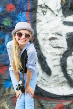 Portrait of smiling hipster girl with retro photo camera against urban wall outdoors