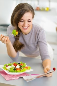 Happy young housewife eating fresh salad and reading magazine in kitchen