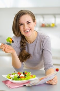 Smiling young housewife eating fresh salad and reading magazine in kitchen