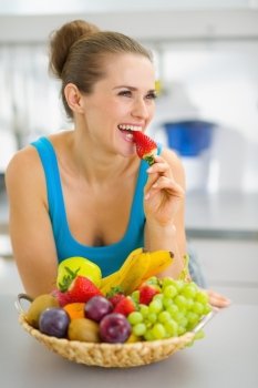 Happy young woman eating strawberry in modern kitchen
