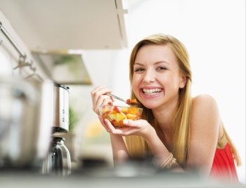 Smiling young woman eating fresh fruits salad in kitchen