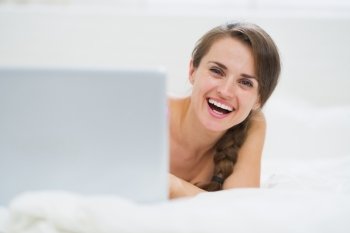 Smiling young woman looking out from laptop while laying in bed