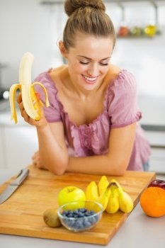 Happy young housewife eating banana in kitchen