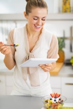 Smiling young housewife eating fruits salad and using tablet pc