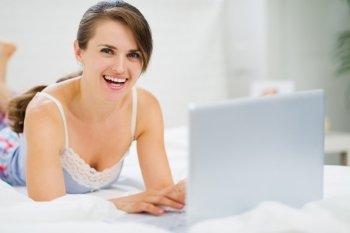 Smiling young woman laying on bed and using laptop