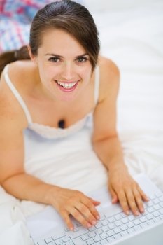 Smiling woman laying on bed and working on laptop. Upper view