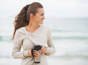Happy young woman in sweater on beach with cup of hot beverage looking on copy space