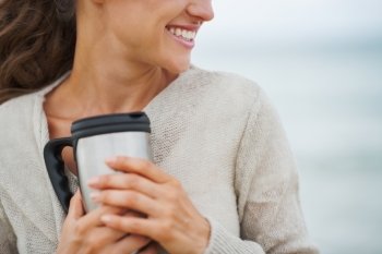 Closeup on happy young woman in sweater on beach with cup of hot beverage