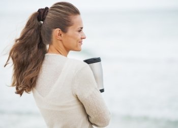 Happy young woman in sweater on beach with cup of hot beverage . rear view