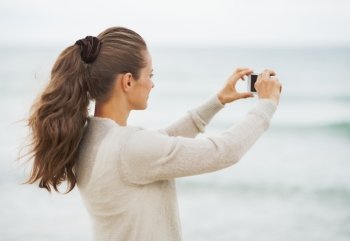 Young woman in sweater on beach taking photo using cell phone
