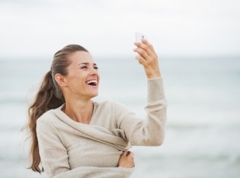 Smiling young woman in sweater on beach taking self photo using cell phone