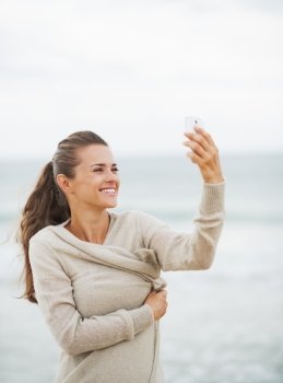 Happy young woman in sweater on beach taking self photo using cell phone