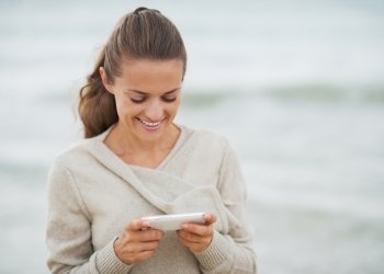 Smiling young woman in sweater on beach writing sms