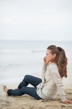 Happy young woman in sweater sitting on lonely beach and talking cell phone