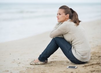 Young woman in sweater with cell phone sitting on lonely beach and looking into distance