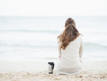 Cup of hot beverage near young woman in sweater sitting on lonely beach . rear view