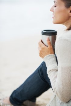Young woman in sweater sitting with cup of hot beverage and relaxing on lonely beach
