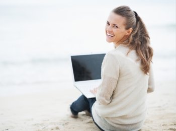 Smiling young woman in sweater sitting on lonely beach and using laptop