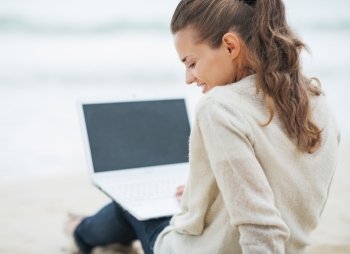 Happy young woman in sweater sitting on lonely beach and using laptop