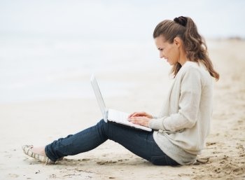 Young woman in sweater sitting on lonely beach and using laptop