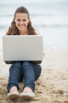 Smiling young woman in sweater sitting on lonely beach with laptop