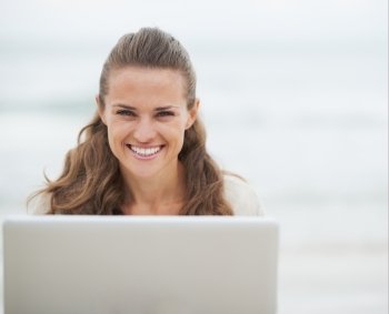 Portrait of smiling young woman in sweater sitting on beach with laptop