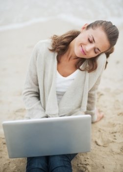Relaxed young woman in sweater sitting on lonely beach with laptop