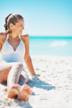 Smiling young woman in swimsuit sitting on beach and looking on copy space