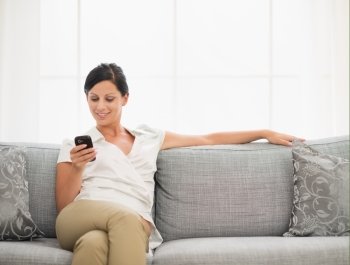 Happy young woman sitting on sofa and reading sms on mobile phone