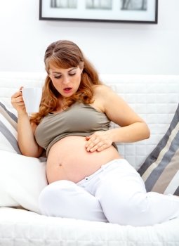 Surprised beautiful pregnant woman sitting on sofa at home and  holding cup of tea in hand
