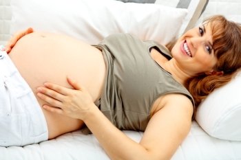 Smiling beautiful pregnant female lying on sofa at home and  holding her belly.
