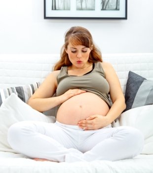 Beautiful pregnant woman sitting at home on sofa and blowing kiss her belly.
