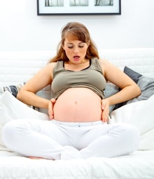 Surprised  beautiful pregnant woman sitting on sofa at home and holding her belly.
