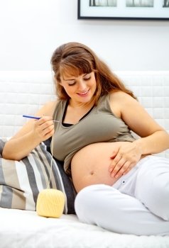 Smiling beautiful pregnant woman sitting on sofa and knitting for her baby.

