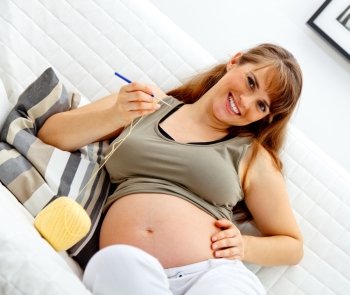 Smiling beautiful pregnant  female sitting on sofa and knitting for her baby.
