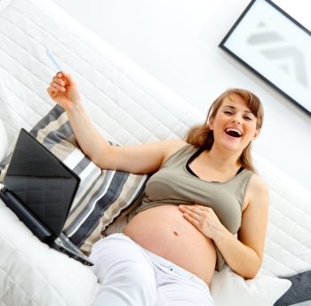 Laughing beautiful pregnant woman sitting on sofa at home with laptop and credit card
