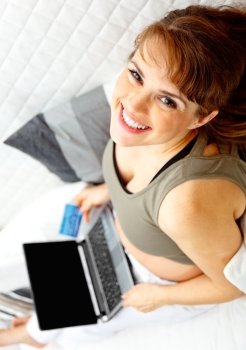 Smiling beautiful pregnant female on sofa at home with the laptop and credit card.
