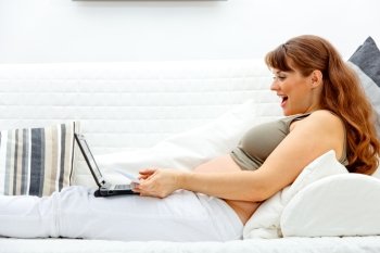 Pleased beautiful pregnant woman sitting on sofa at home with laptop and credit card
