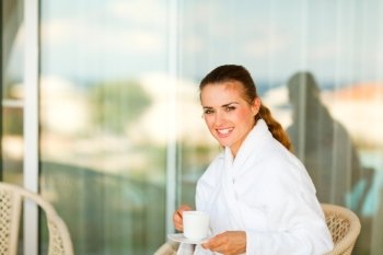 Smiling pretty woman in bathrobe sitting at table on terrace and having cup of tea
