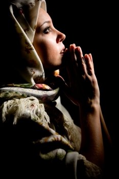 Portrait of beautiful praying girl with old russian shawl on head on black background. 
