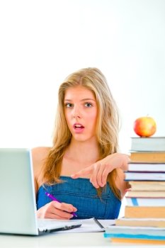 Curious young girl sitting at table with books and pointing on laptop 
