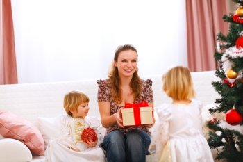 Two twins girl presenting gift to mother near Christmas tree

