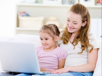 Young mother with little daughter sitting together at home and looking at laptop 