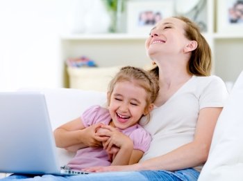 Laughing happy young mother and little daughter with laptop on the sofa at home