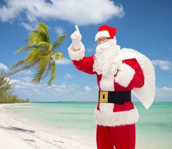 christmas, holidays, gesture and people concept - man in costume of santa claus with bag pointing finger up over tropical beach background