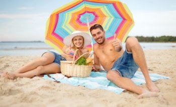 summer, holidays, vacation and happy people concept - smiling couple lying on the beach under colorful umbrella and showing thumbs up